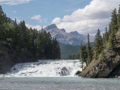 A wide, turbulent waterfall cascading over a rocky ledge into a turquoise river, framed by dense evergreen forests and steep rocky banks, with a prominent mountain peak visible in the background under a partly cloudy sky.