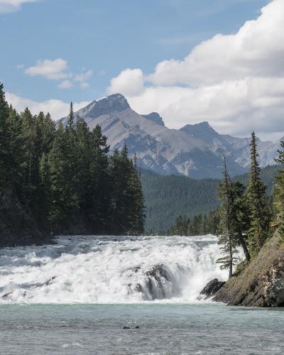 A wide, turbulent waterfall cascading over a rocky ledge into a turquoise river, framed by dense evergreen forests and steep rocky banks, with a prominent mountain peak visible in the background under a partly cloudy sky.