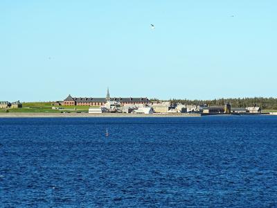 A wide shot of a reconstructed 18th-century colonial settlement with stone walls, red-roofed buildings, and a church steeple situated on a grassy peninsula across a body of blue water.