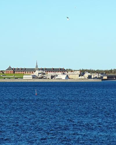 A wide shot of a reconstructed 18th-century colonial settlement with stone walls, red-roofed buildings, and a church steeple situated on a grassy peninsula across a body of blue water.