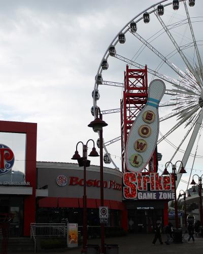 The image shows a street-level view of an entertainment district featuring a large white Ferris wheel, a "Boston Pizza" restaurant, and a "Strike Zone" bowling arcade sign.