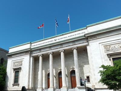 The image shows the neoclassical facade of a large stone building with four prominent columns, featuring the text "MUSÉE DES BEAUX-ARTS DE MONTRÉAL M THE MONTREAL MUSEUM OF FINE ARTS" inscribed on the frieze, with three flags flying above.