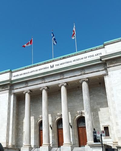The image shows the neoclassical facade of a large stone building with four prominent columns, featuring the text "MUSÉE DES BEAUX-ARTS DE MONTRÉAL M THE MONTREAL MUSEUM OF FINE ARTS" inscribed on the frieze, with three flags flying above.