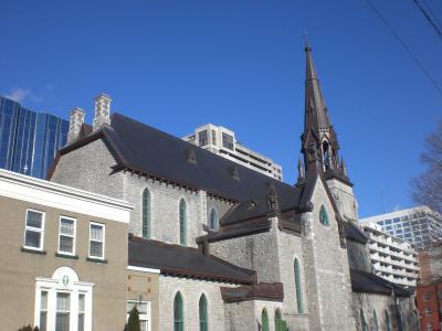 A stone Gothic Revival church with a prominent central spire and green-trimmed arched windows, viewed from the side against a blue sky with modern buildings in the background.