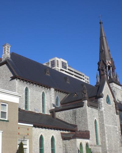 A stone Gothic Revival church with a prominent central spire and green-trimmed arched windows, viewed from the side against a blue sky with modern buildings in the background.