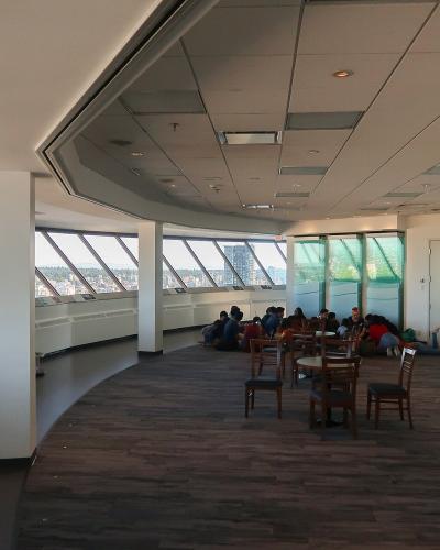 An interior view of a curved observation deck with large angled windows overlooking a city and water, featuring visitors sitting on the floor and at tables.