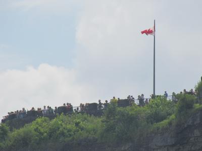A crowd of people standing along a stone viewing wall on a cliff edge, with a Canadian flag flying on a tall pole against a cloudy sky.