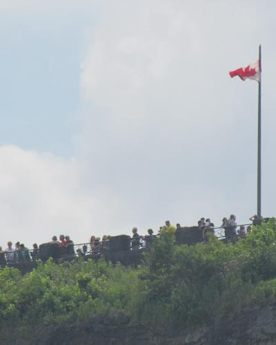 A crowd of people standing along a stone viewing wall on a cliff edge, with a Canadian flag flying on a tall pole against a cloudy sky.