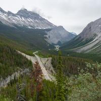 A high-angle view of a paved highway winding through a mountain valley with snow-dusted peaks and coniferous forests, taken from an elevated viewpoint.