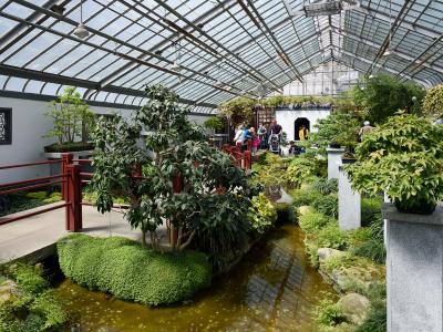 An indoor Chinese-style garden featuring a glass greenhouse roof, a small stream, red wooden railings, manicured greenery, and visitors walking on pathways.