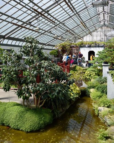 An indoor Chinese-style garden featuring a glass greenhouse roof, a small stream, red wooden railings, manicured greenery, and visitors walking on pathways.